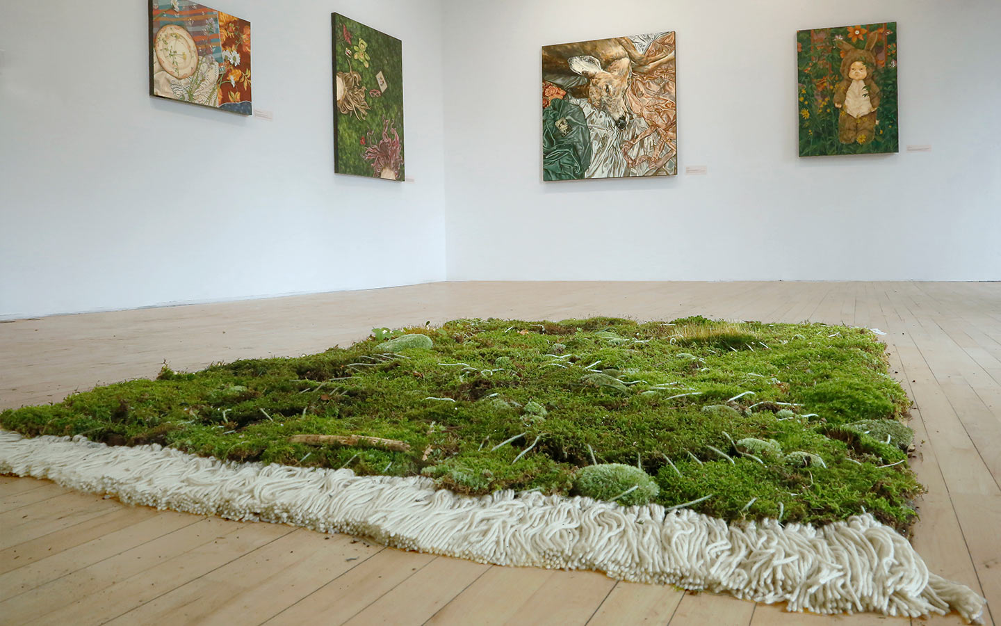 A green mossy like rug in the middle of a white walled gallery space with paintings on the wall.
