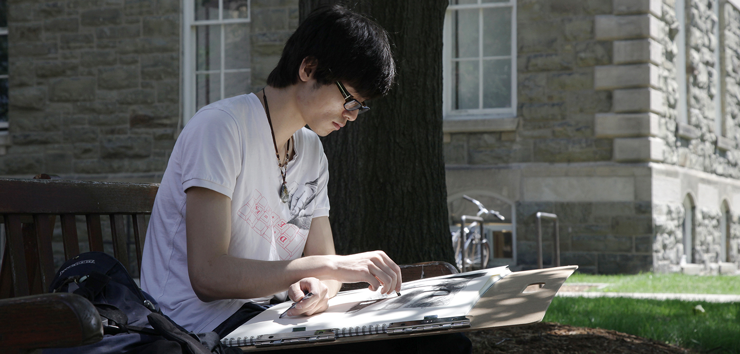 A young man seated outside in dappled sunlight drawing on a board in his lap, a stone building in the background