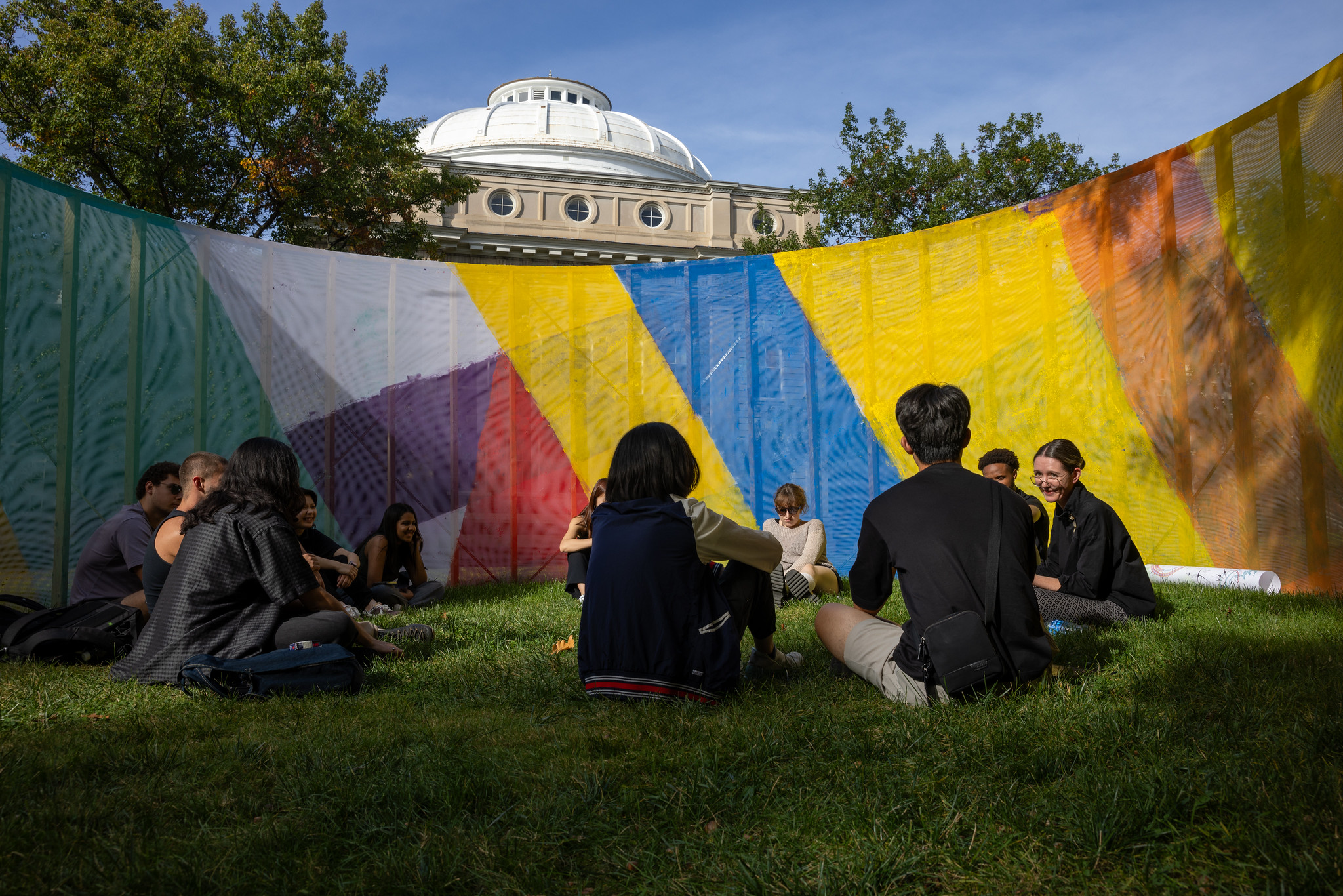 People sitting outside on the grass surrounded by a brightly colored wall.