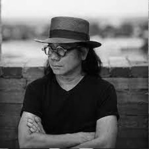 Man with long dark hair wearing glasses, a hat, and a black tshirt