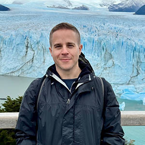 A man dressed in a dark blue jacket smiles at the camera. Behind him is a blue glacier.