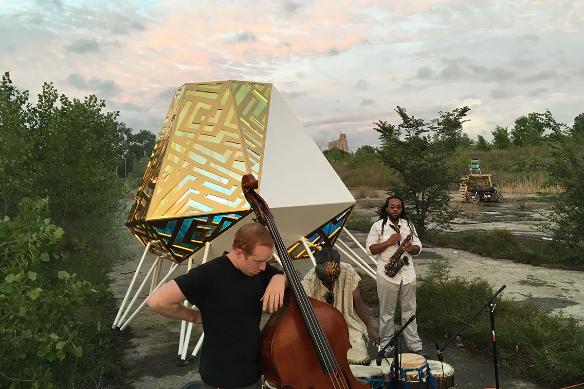Bass, saxophone, and percussion musicians gathered outside in an overgrown lot.