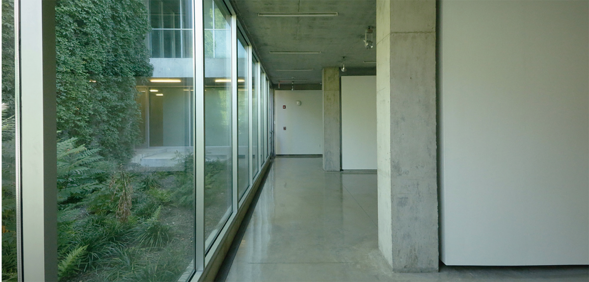 View of the gallery with a glass wall on the north side showing a garden on the outside and a concrete pillar on the right with a dividing wall.
