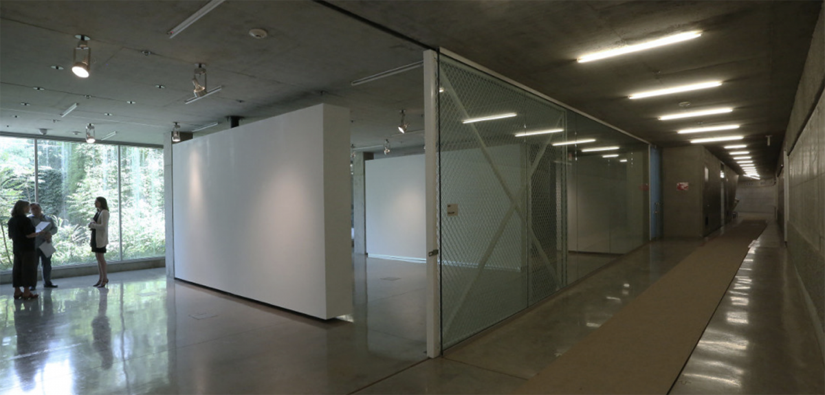 View into a gallery space that has a glass wall on the south entrance side and three people standing on the opposite side of the room.
