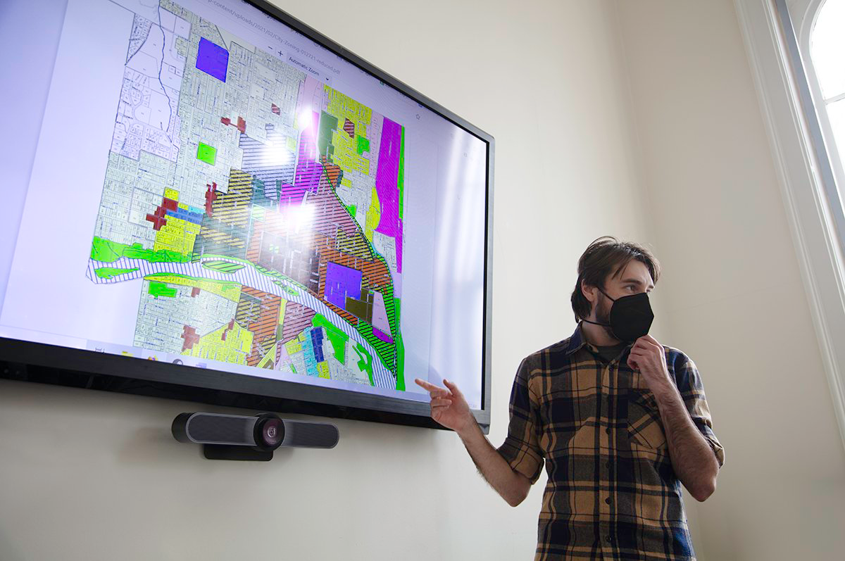 Person in plaid shirt gestures at colorful zoning map displayed on wall TV screen