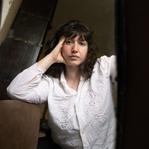 A woman with light skin, brown wavy hair with bangs, and a gold nose ring, wearing a white long sleeved button down shirt and resting her head against her hand as she sits leaning against a couch.