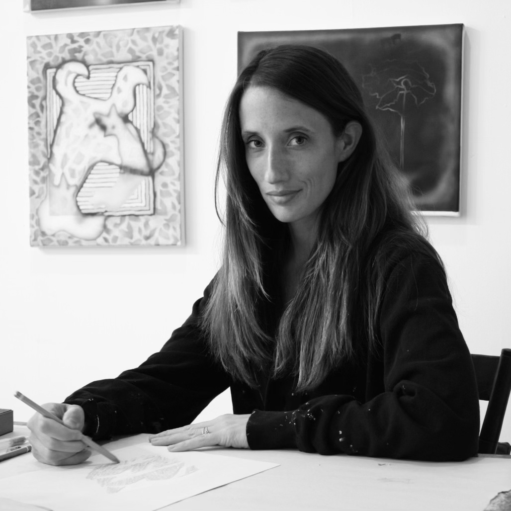 A black and white photo of a woman with long hair, wearing a long sleeved black shirt, sitting at a table and working on a drawing as she looks at the camera. On the wall behind her are two paintings.