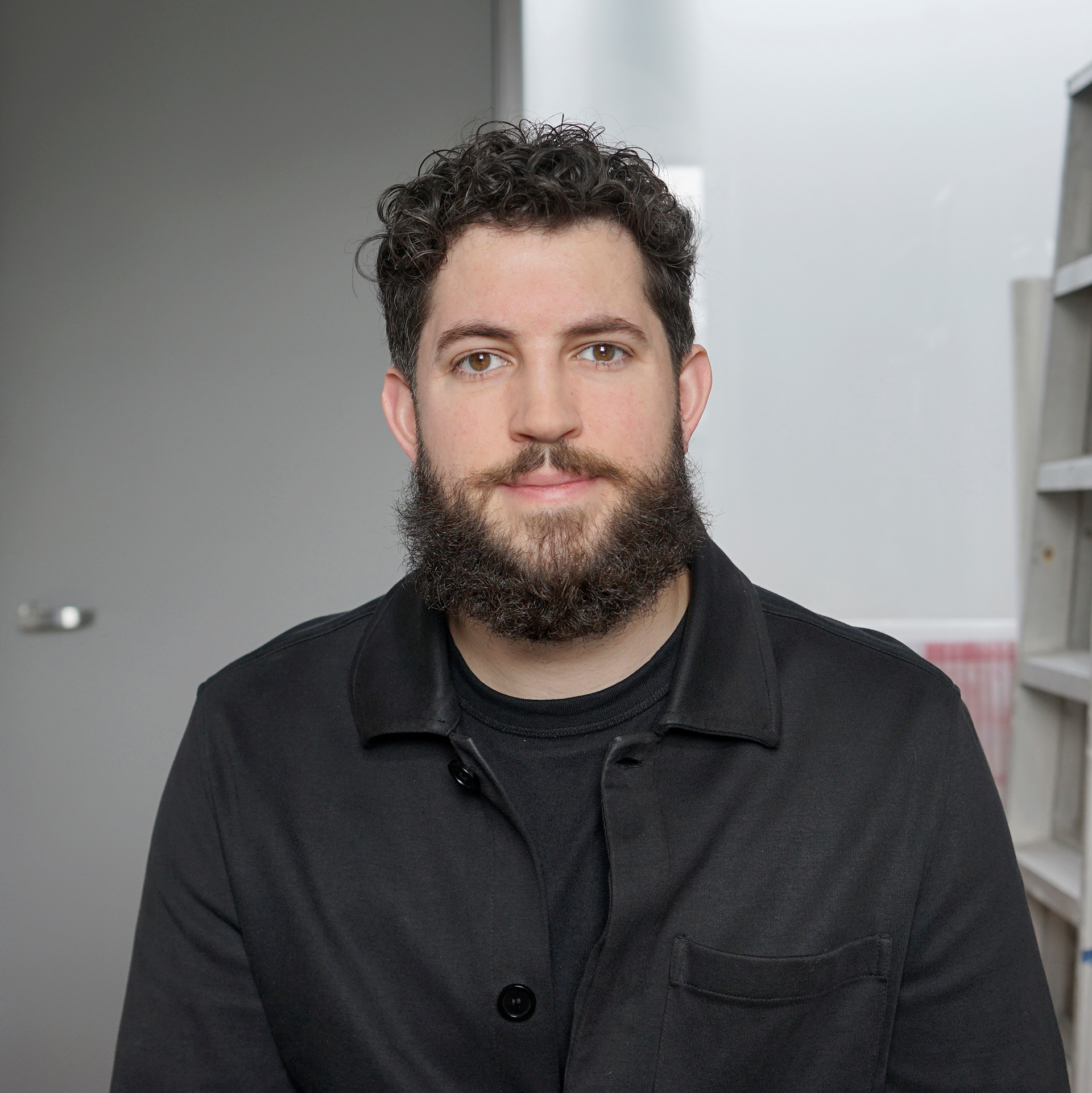 A man with dark curly hair and a dark beard looking at the camera in front of a grey wall