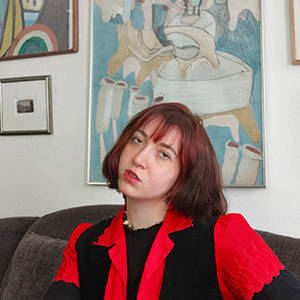 Woman with short reddish brown hair wearing a red and black shirt in front of paintings hanging on the wall.