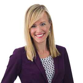 Smiling woman with long hair wearing a purple blazer in front of a white background. 