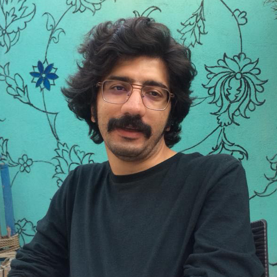 A person with a mustache, glasses, and dark hair wearing a black shirt in front of a teal flowery wall.