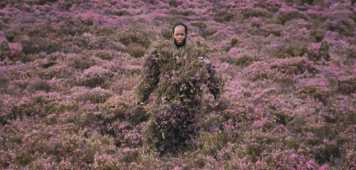 Man covered in dark green and purple grass and flowers with just his head visible.