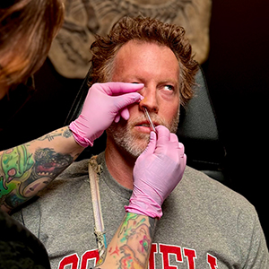 A man with light skin and wavy brown hair and a beard wearing a gray tshirt with red lettering and getting his nose pierced.