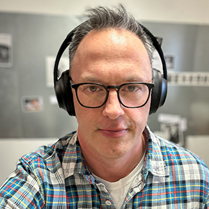 A balding man with light skin and gray hair, wearing black rimmed glasses, black headphones over his ears, and a blue plaid button down shirt over a white t-shirt.