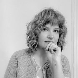 A black and white photo of a woman with light skin, shoulder-length wavy hair with bangs, and a septum piercing, wearing a cardigan and resting her chin on her hand as she looks at the camera.