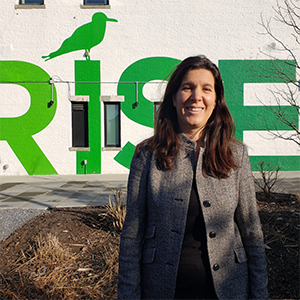 A woman with long brown hair smiles in front of a white building with RISE printed on it in green letters. The outline of a bird is painted on top of the I in RISE.