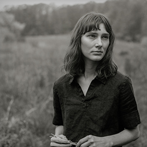 A black and white photo of a person with light skin and loosely wavy hair with bangs, wearing a short-sleeved button-down shirt and looking off into the distance as they stand in a field.