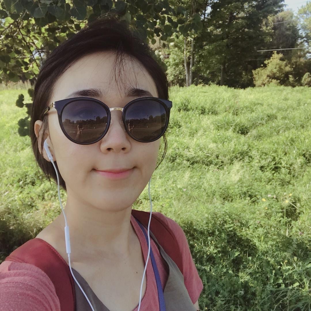 A person with short dark hair wearing sunglasses and headphones with grass and trees in the background.