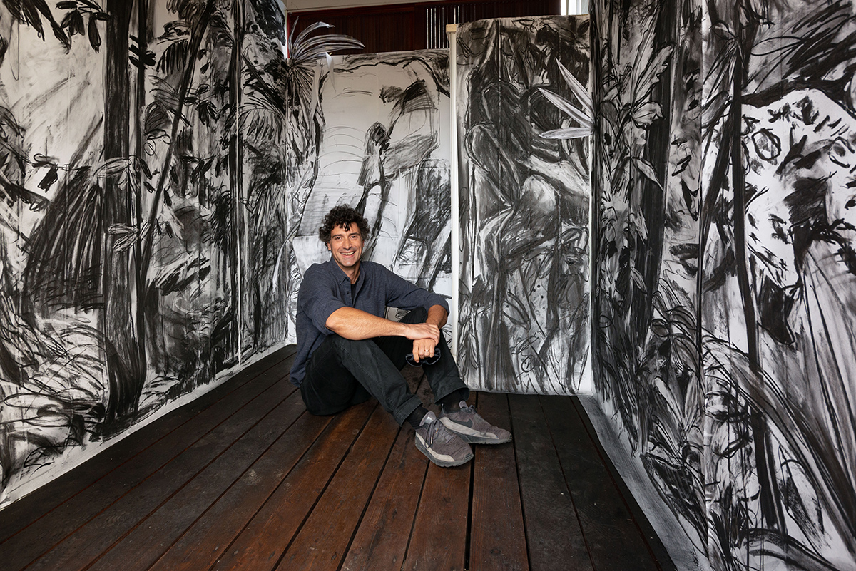 man sitting in a room with wall drawings in charcoal of trees and leaves