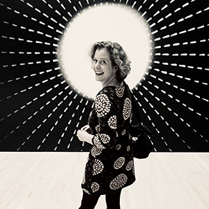 Black and white view of a person with light skin, chin length curly hair wearing an abstract patterned dress, smiling as they look behind their shoulder in front of a black and white sun-like decorative background.