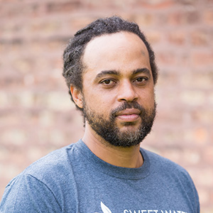 person with beard in a blue shirt looking at the camera against a brick background