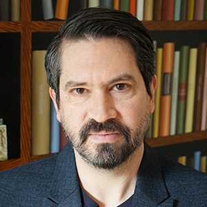 A person in front of a bookshelf with a suit jacket and a beard.