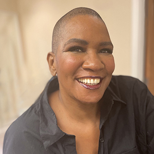 A Black woman with closely shaved dark hair, wearing a dark gray button-down shirt, and smiling.