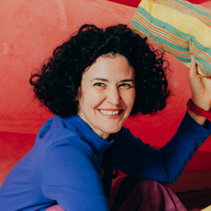 A smiling person with dark curly hair, wearing a long-sleeved blue shirt