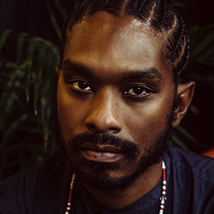 A man with a beard and Afro-textured hair worn in cornrows, wearing a dark blue shirt and a red and white necklace.