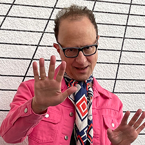 A man with brown hair and black square glasses, wearing a pink jacket and multi-colored patterned scarf, against a black and white grid background. He is holding his hands up as he looks at the camera.