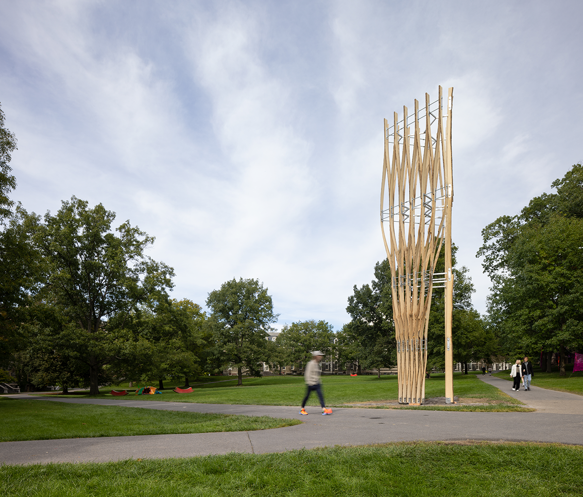 Tall wooden sculpture on a green lawn with people passing