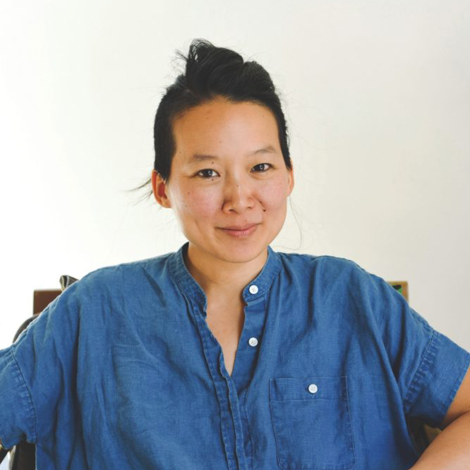A person with black hair and a blue button down shirt with their arms on their hips.
