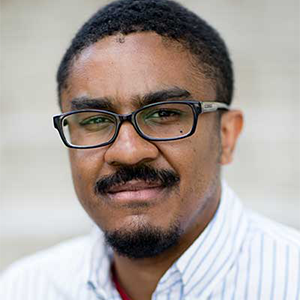 Black man with a dark moustache and beard wearing glasses and a striped white button down shirt