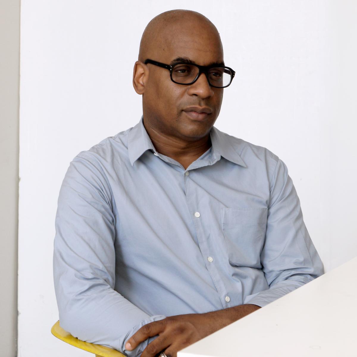 A photo of a dark-skinned bald man in three quarter profile. He is wearing a light blue button down shirt and black frame glasses.