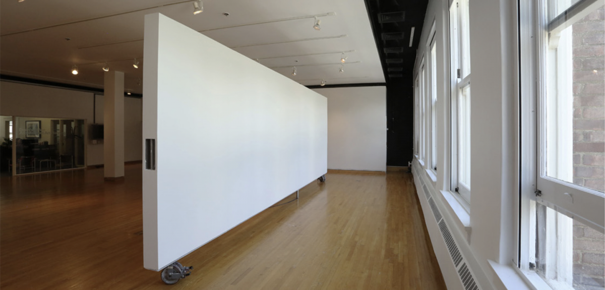 Room with white walls and wood floor, a moveable wall in the center, glass office wall on left and wall of windows on right.