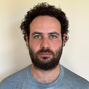 Person with light skin, short curly hair and brown beard in a grey t-shirt looks unsmiling at the viewer in front of a cream background.