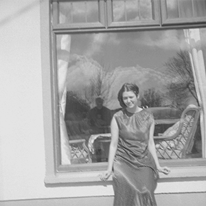 A black and white photo of a woman with light skin and dark hair, wearing a full-length dress and leaning against the window of a house.