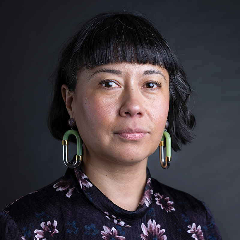 Woman with dark chin-length hair and bangs wearing a floral shirt with large dangling earrings.