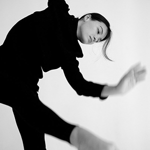 Black and grey view of a Japanese-Taiwanese American person with dark hair in a ponytail wearing black dancing in front of a white background. 