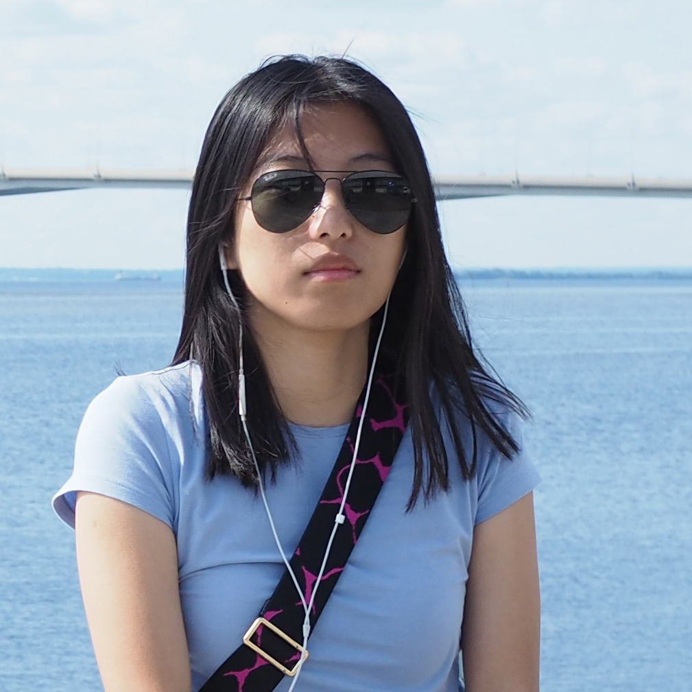 A person with dark hair and sunglasses with earbud headphones in standing in front of water.
