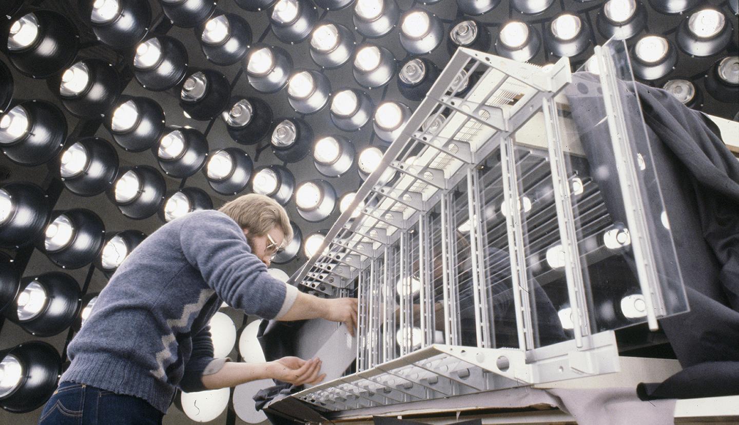A person adjusting a glass window on an architectural model surrounded by large light bulbs.