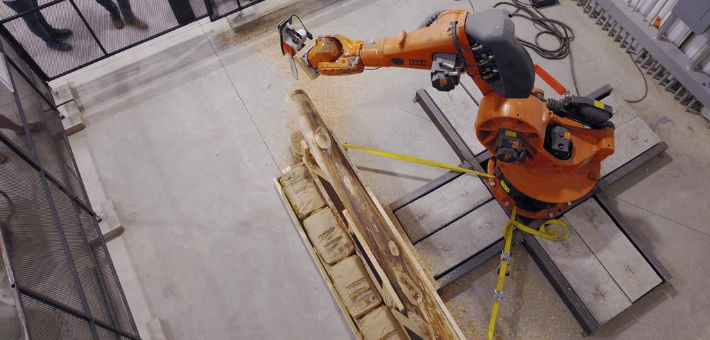 aerial view of an orange and black robot arm cutting a wooden log in a concrete-floored studio