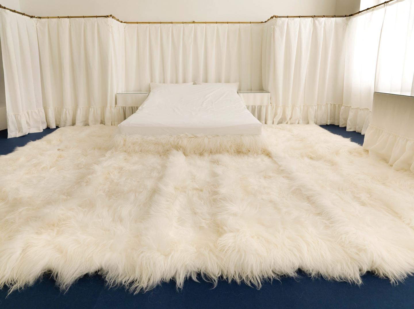 A room with cream colored drapes on the walls and a furry rug and a white bed on blue carpet.
