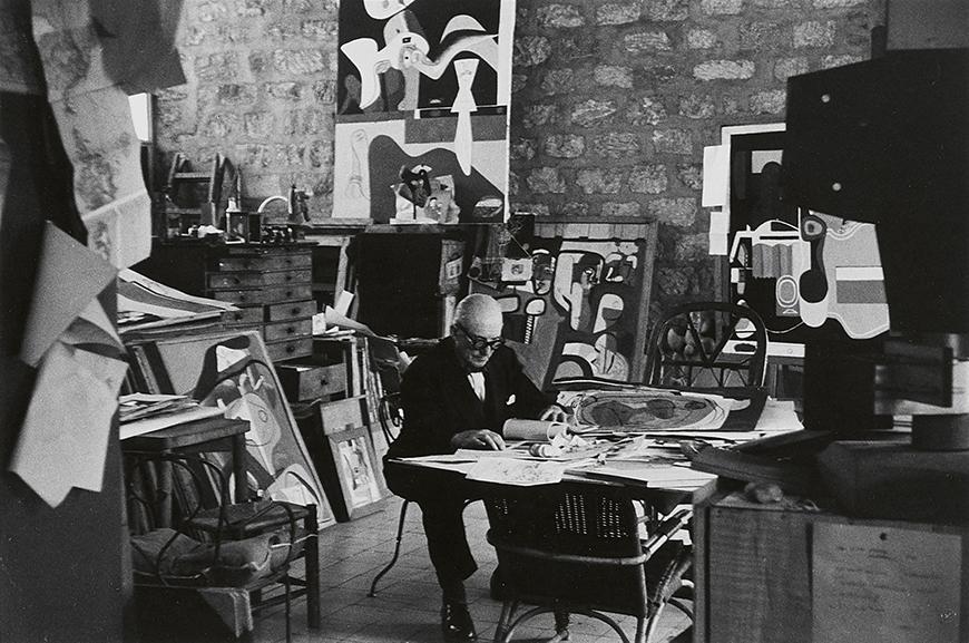 black and white image of a man seated at a desk in a studio with abstract art behind him