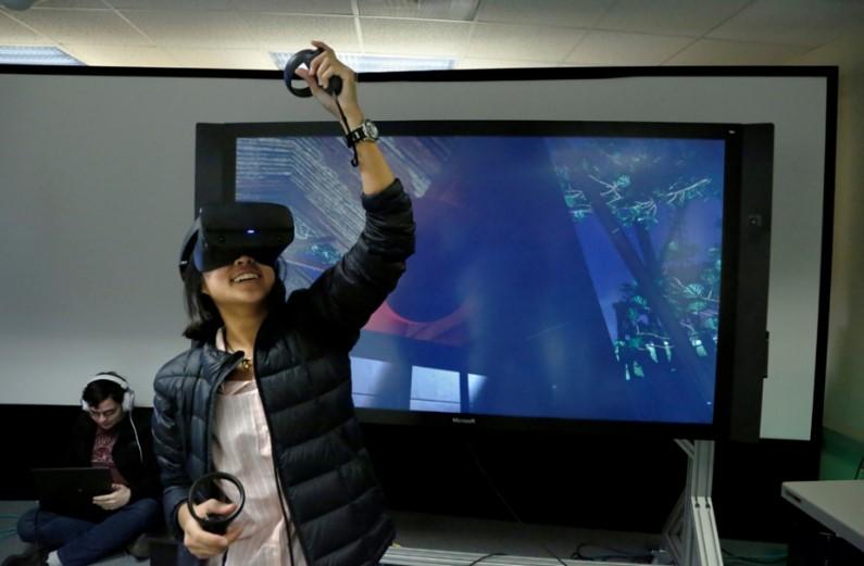 A person wearing a virtual reality headset and holding virtual reality remotes in front of a large screen.