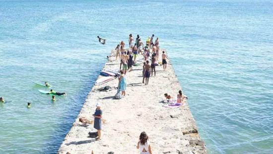 People standing on a rock or cement jetty in the ocean with people playing and swimming.