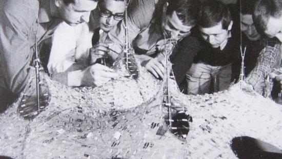 Black and white photo from the 70s of six men looking closely at a surface