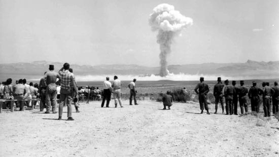Crowd of people on a flat sandy area, a mushroom cloud rises in the sky.