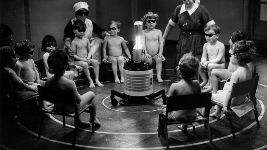 naked children and nurses wearing sun glasses gathered around a UV light