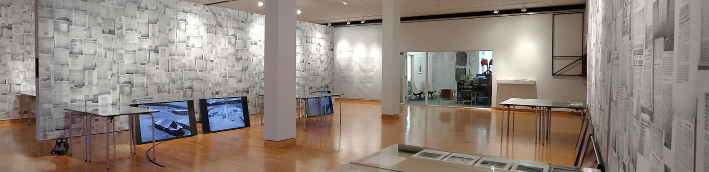 A white walled gallery with pieces of paper on the walls with television screens sitting on the floor.